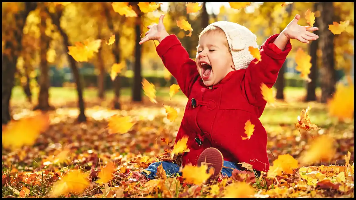 Child in a red coat raises both arms joyfully as golden autumn leaves fall around them.