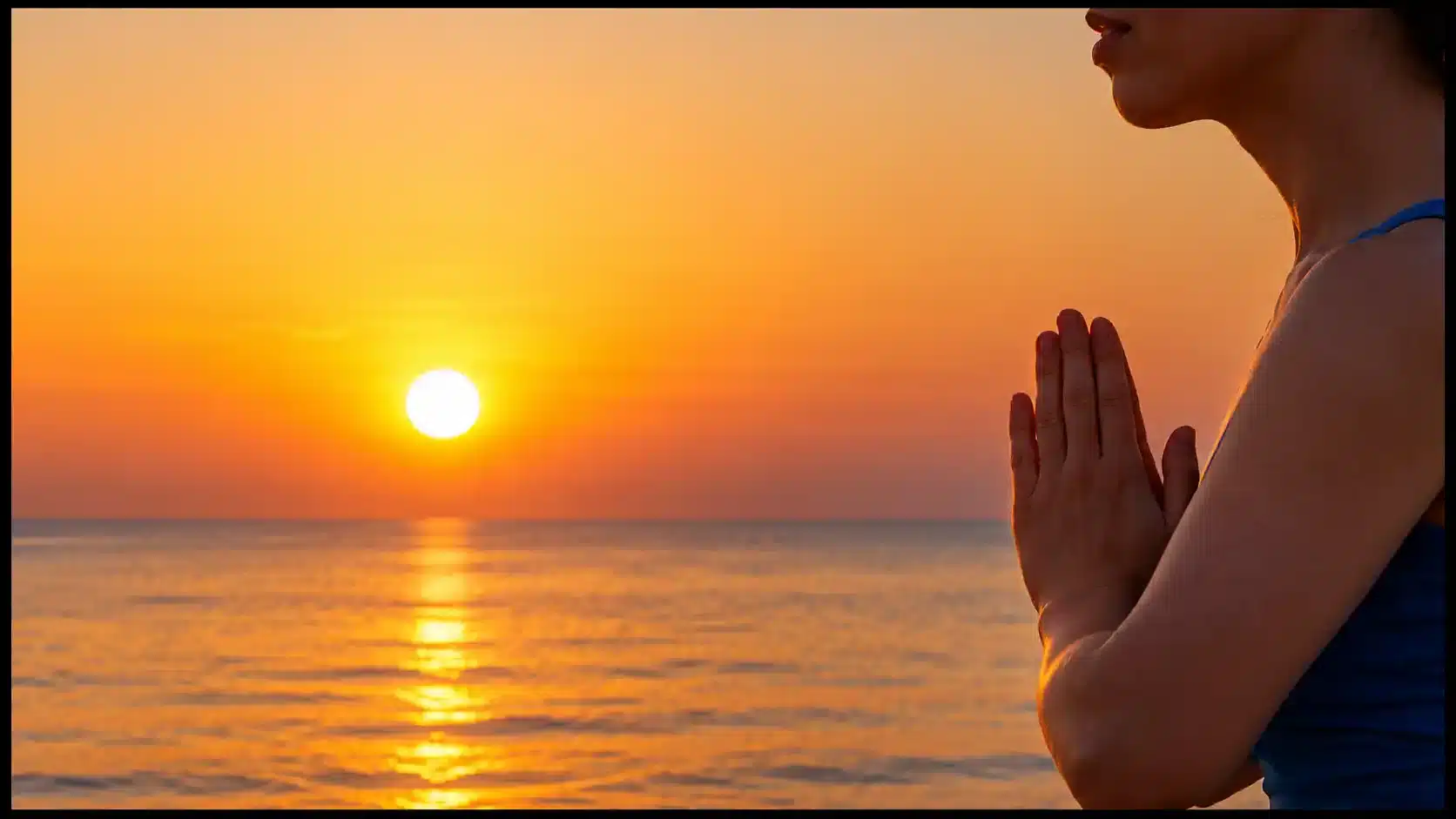 A person holds their palms together at chest level beside calm water under an orange sunset.
